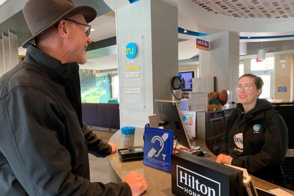 A man in a hat and glasses talks to a desk agent at the counter. A hearing loop sign and Hilton Honors sign on the counter.