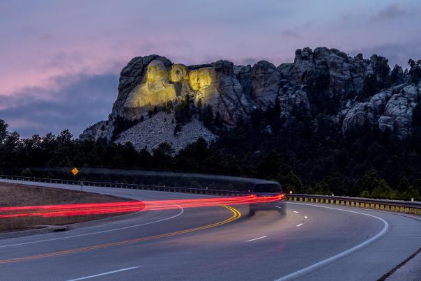Mount Rushmore, lit up by yellow floodlights at dusk, sits in the background as a vehicle traveling down the highway is trailed by a long line of red taillights, created by a long-exposure photo.