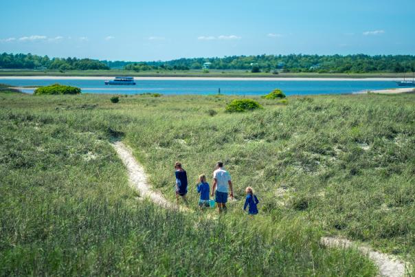 Family of four walking on a path on Masonboro Island with the Carolina Runner sailing in the background.