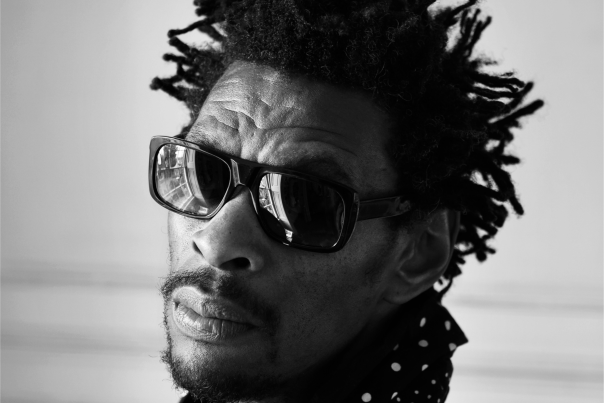 A black and white photo of Daddy G - a British DJ and founder of Massive Attack. Daddy G has black hair and wears black sunglasses. The photo is of his head and shoulders and he wears a polka dot scarf. He looks towards the camera without smiling and stands in front of a white wall.