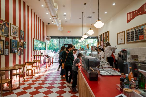 The interior of OddFellows' Ice Cream Co. features an ice cream counter stretching away from the camera. The left wall is red and white striped with classic paintings of odd or novelty subjects (a toy monkey with symballs, a seal in a hot dog cart, etc.). The floor is checkered with red and white tiles, set with a few small, round, 2-person tables and a bar with four stools. There are several families with 5- to 12-year-old children peering at the ice cream flavors.