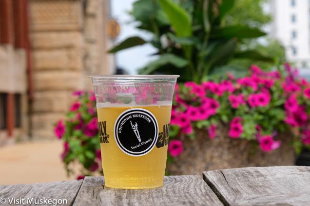 plastic cup of beer sits on picnic table in downtown muskegon. a circular black sticker reads social district. hot pink flowers and green foliage are in the background.