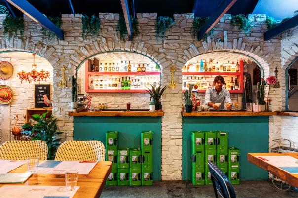 A vibrant Mexican-inspired bar with stone archways, colourful decor, and a bartender crafting drinks behind a wooden counter.