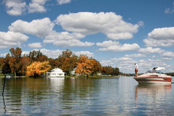 A man on stands on the front a red and white boat on Silver Lake in Fenton, Michigan with trees turning colors in the background.