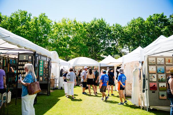 A wide-angle view of The Woodlands Waterway Arts Festival at Town Green Park. Visible is a winding row of white pop-up tents layered with colorful paintings, photos, jewelry, and other works of fine art. Visitors mill about in and among the tents, admiring the displays. Behind everything are bright green trees and a bright blue sky.