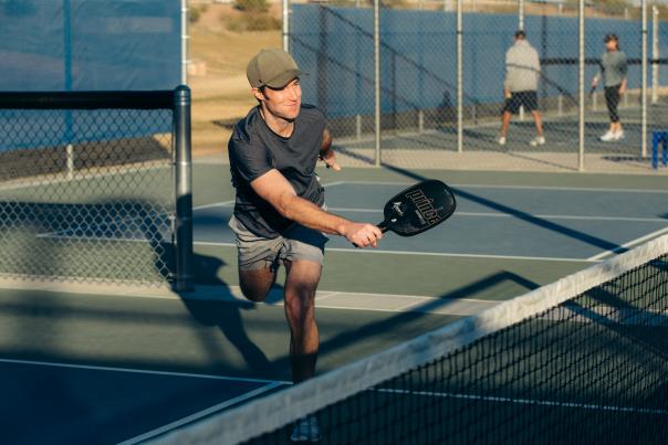 man is green baseball cap, dark tee shirt and grey short is playing pickleball on sunny outdoor court