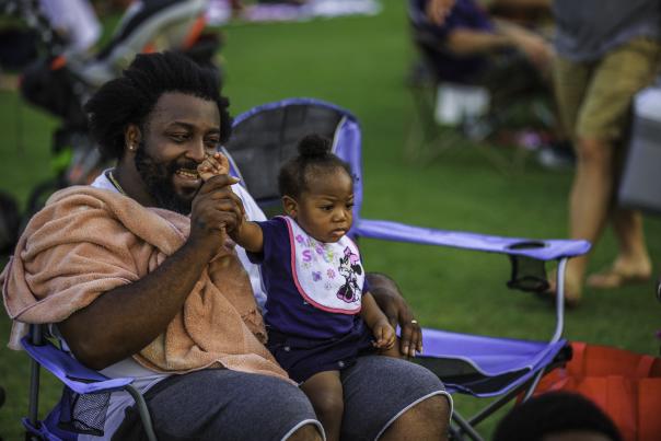 Father and child sitting together at Festival Park during a Fourth of July celebration, watching the event with other attendees nearby.