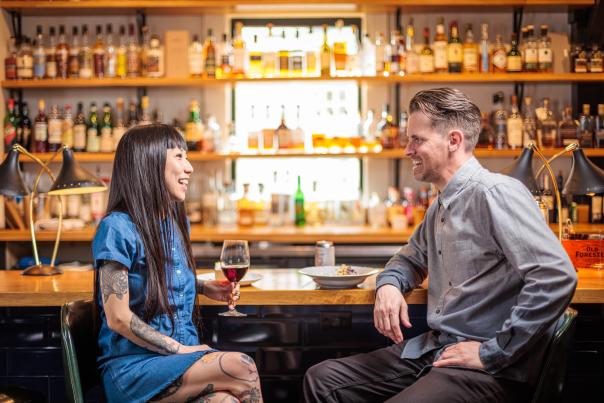 A couple of people sit at a bar, each with a drink. They are smiling.