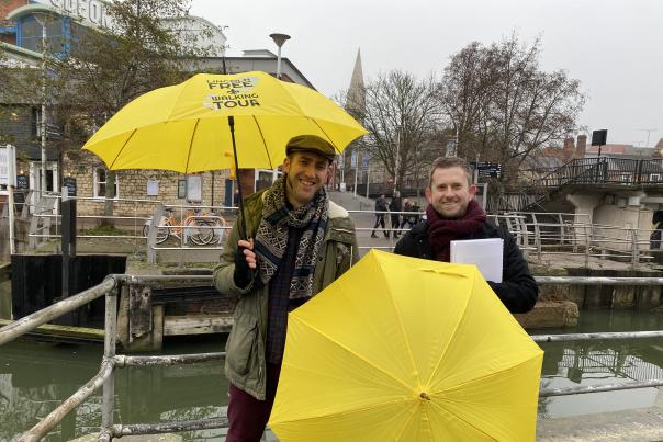 two people stood by a river holding yellow umbrellas and smiling at the camera