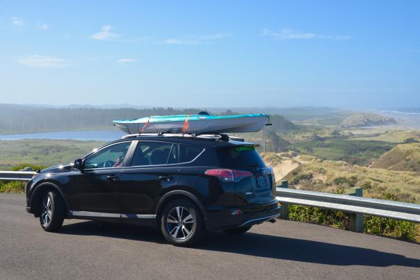 Car with a kayak on the roof is parked with dunes, a lake and the ocean in the background.