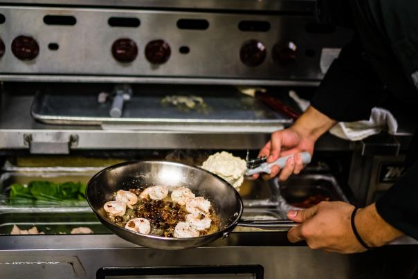 A pair of hands holds out a pan full of sizzling shrimp over an oven while scooping another ingredient into the pan