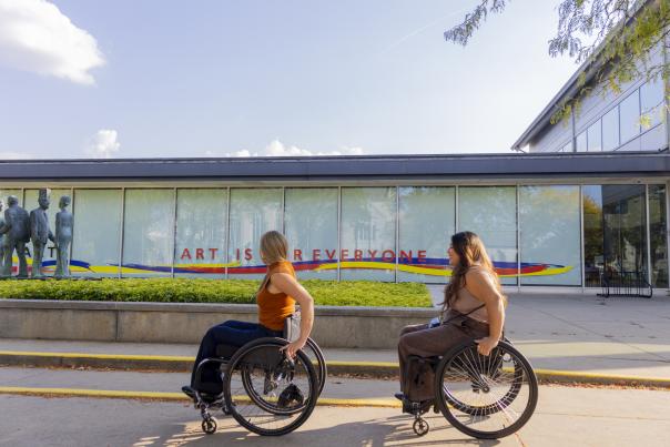 Two young women in wheelchairs roll on the sidewalk outside of the Kalamazoo Institute of Arts' sign stating "Art is for Everyone."