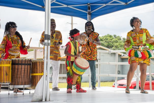 Five people dressed in brightly colored clothes, honoring their ethnicity, playing various percussion instruments on an outdoor stage, under a blue canopy.