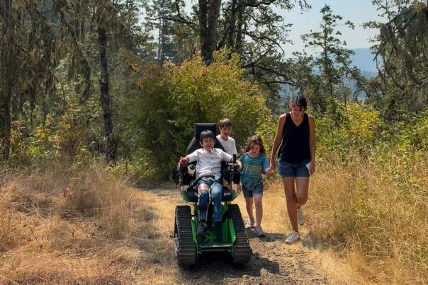 A family walks and rolls using a track chair along a golden path in the forest.