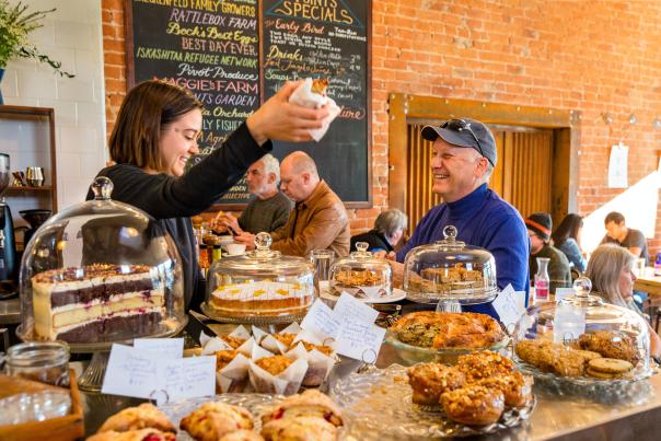 woman standing behind a counter, filled with breakfast goods and pastries, handing a pastry to a smiling older man in line