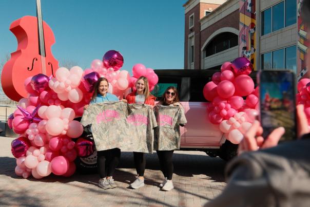 three people stand in front of a pink car and balloons outside of a Megan Moroney concert.