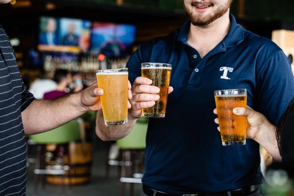 Two men and a woman (their faces out of the frame) each hold a glass of beer in front of them while they talk at Kirby Ice House.
