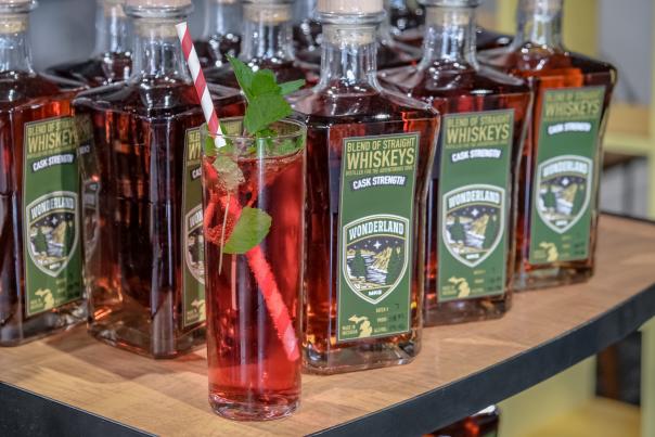 Bottles of Wonderland Distilling whiskey sit on shelf behind tall red cocktail trimmed with green mint leaves and red and white striped straw