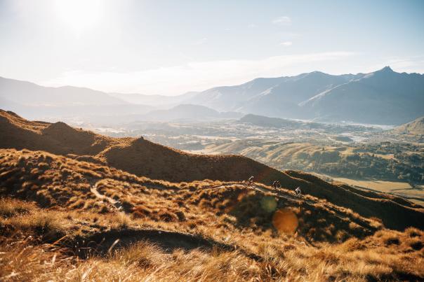 Far shot of three bikers descending a golden tussock-covered mountain with valley and mountain in the background