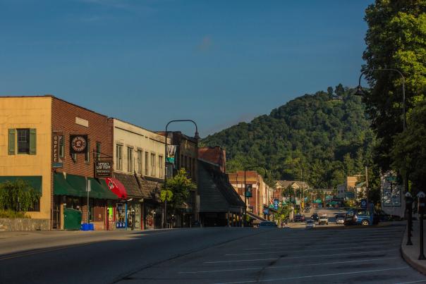 A nearly empty street with historic-looking buildings on the left side. There is a tree covered mountain rising in the background.