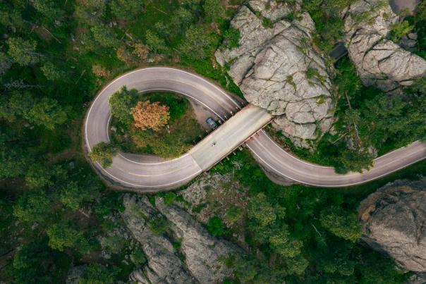 Aerial view of Needles Highway forming a figure-eight, surrounded by lush green trees and large gray rocks. A bridge spans a narrow gap, creating a serene landscape.