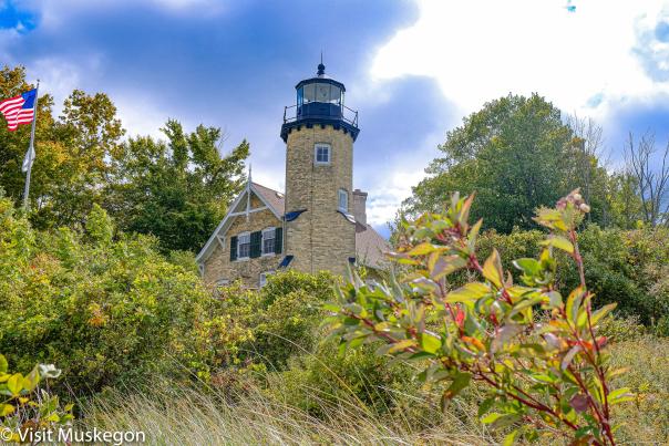 historic brick lighthouse sits atop dune grass and shrub covered hill below blue sky. an american flag flies to the left.