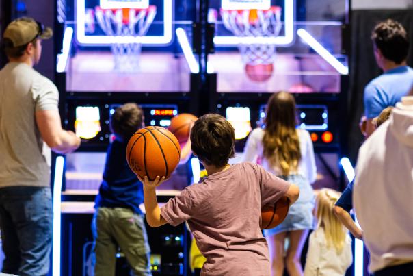 The back of a child tossing a basketball at a arcade backstop.