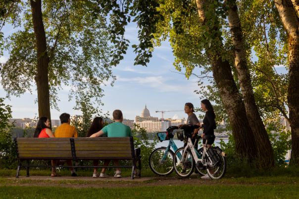 A shot of the city skyline over the lake while a group of people sit on a bench and two others stand next to them with bikes overlooking the views.