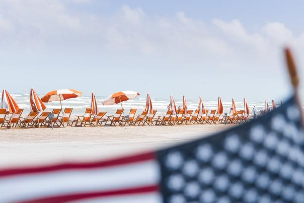 In focus is the background of the photo, which shows a row of orange and white beach chairs and umbrellas on the beach. An American flag is in the foreground, blurred.