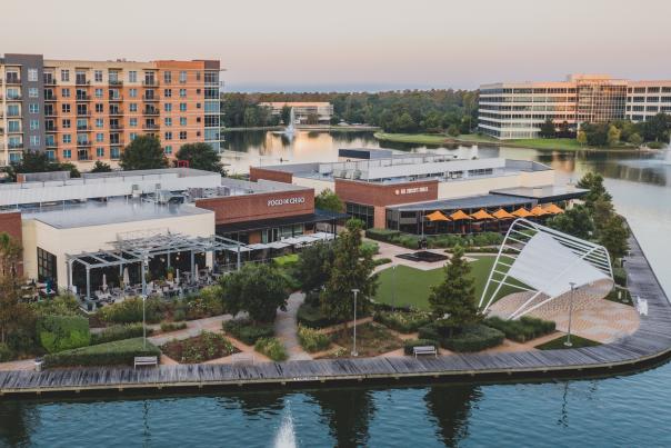 An aerial photo of Hughes Landing. Lake Woodlands wraps around the bottom, righthand side, and toward the horizon of the frame. On the land in the center is Restaurant Row, featuring a building with no visible name, Fogo de Chao, and Del Frisco's Grille. Along the dock is the Hughes Landing Band Shell, and behind the restaurants is a large apartment building. On the opposite shore (back right of the frame) are lots of trees and two large office buildings. A large fountain is visible in the lake in the back-middle part of the frame.