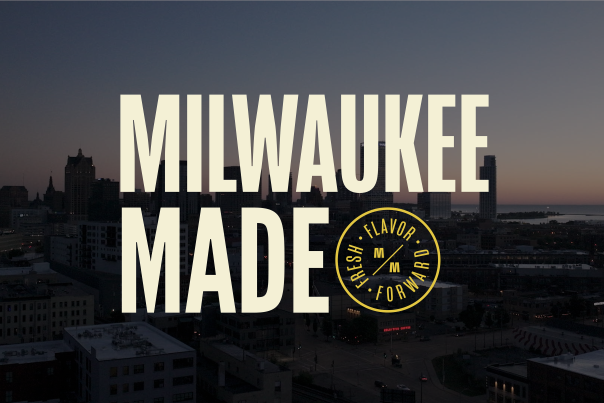 Aerial view of Milwaukee's downtown skyline at dusk with tall buildings silhouetted against a gradient sky. Large cream-colored text reads 'MILWAUKEE MADE' overlaid on the cityscape, with a circular yellow badge logo containing 'FRESH FLAVOR FORWARD' and stylized 'MM' letters positioned to the right of 'MADE'.