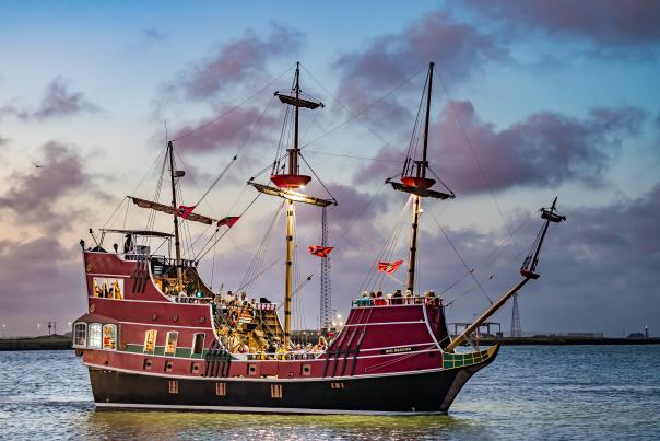 Large red pirate ship in the ocean with lights on in facing the deck. The sun has almost set and gives the sky and clouds a purple tint.