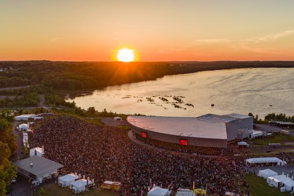Aerial shot of Lakeview Amphitheater, large crowd infront of amphitheater set on the shores of Onondaga Lake, a fiery orange sunset can be seen in the horizon