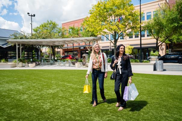 Two women dressed for fall walk through Central Park at Market Street. The blonde woman (left) is carrying a small yellow shopping bag from Kendra Scott. The dark-haired woman (right) is carrying larger bags in purple (Hemline) and white (Golden Gray Boutique).