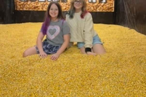 Two girls sit atop thousands and thousands of yellow corn kernels in an enclosed corn pit.