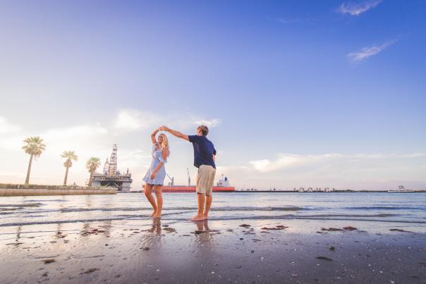 A couple stands on the beach, mid-twirl. In the background a few palm trees and a ship are visible.