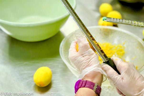 Close-up of gloved hands zesting a yellow lemon over a bowl of yellow shavings; lemons and green bowls sit nearby.