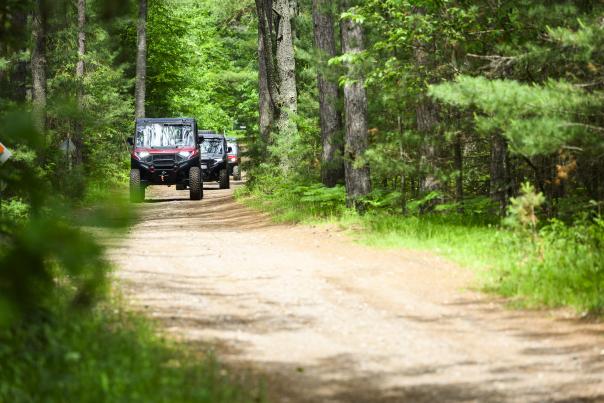 Three UTVs are driving down a dirt trail surrounded by forest.