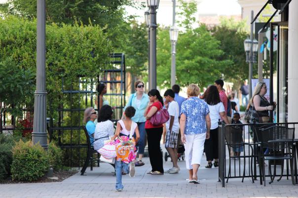 Visitors stroll along the sidewalks at Bridge Street Town Centre, browsing shops and enjoying the outdoor shopping district. Trees, benches, patio seating, and storefronts line the lively pedestrian walkway.