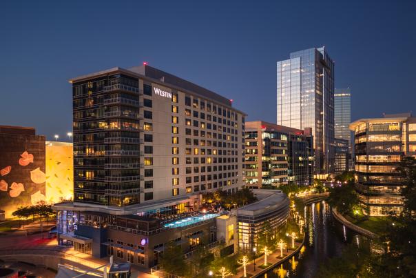 A drone shot of The Westin and The Woodlands Towers at dusk. Surrounding buildings and trees along The Waterway glow with soft golden lights.