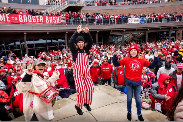 Three women dressed in UW badger gear jumping and cheering with a crowd of Badger fans behind them.