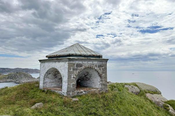 Markøy fyr – et historisk steinhus med buede åpninger og metalltak, plassert på en gresskledd høyde med utsikt over havet og Korshamn skjærgård under en dramatisk skyhimmel