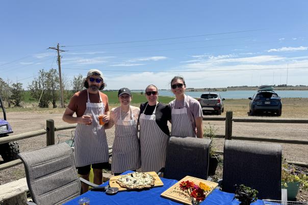 Four people standing on a patio with a lake in the background with food on the table