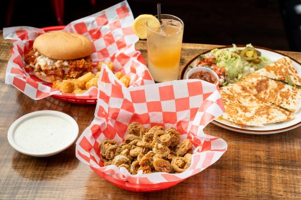 A spread of three Lankford's dishes: a fried chicken sandwich (buffalo flavored) with tater tots, a basket of fried jalapeno bottle caps, and a plate of quesadillas.