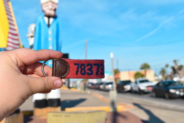 A hand holds up a red needlepoint keychain with the numbers "78373." In the background is Alister in Port A.