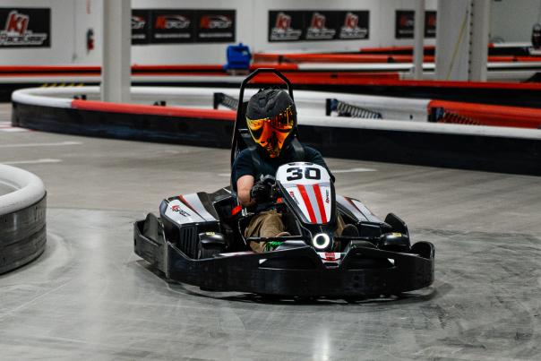 A man turns a go kart around a corner at K1 Speed. He's wearing a dark T-shirt, tan cargo shorts, black racing gloves, and a black motorcycle helmet with a colorful reflective visor. The track is gray and rubbery, the barriers alternating in red and white stripes. K1 posters line the back wall.