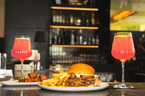 Burger and fries from The 7th Street Standard served alongside two bright pink spritz cocktails at a dimly lit bar.