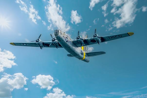 A historic four-engine bomber aircraft flies overhead against a bright blue sky dotted with white clouds. The polished silver plane is captured from below, highlighting its distinctive wingspan and vintage aviation design.