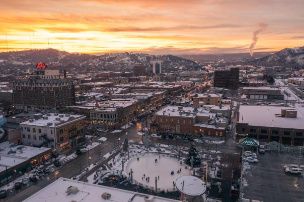 A snowy cityscape at sunset, with warmly lit buildings and streets. A central ice rink with skaters is surrounded by hills and a vibrant orange sky.