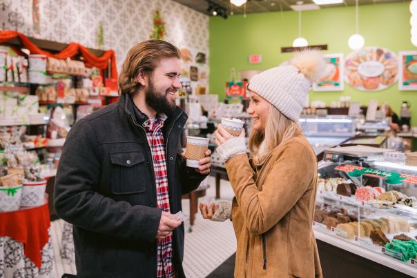A smiling couple enjoys hot drinks inside a festive Kilwins decorated with holiday colors. They stand near a display case filled with assorted sweets and baked goods, creating a warm and cheerful atmosphere.
